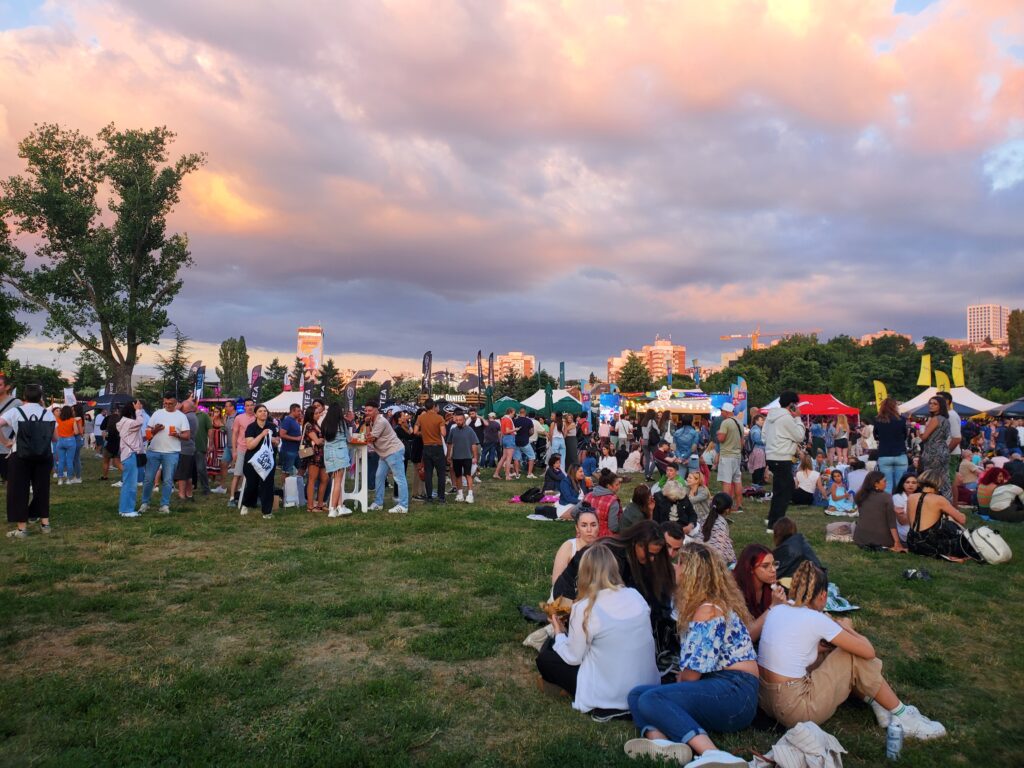 A large crowd sits and stands in a field, with food stands in the background. The sky is pink and purple.