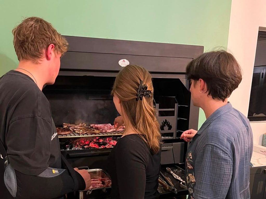 A woman faces a built-in fireplace, adding meat onto the grill, flanked by two men observing her newly acquired skill.