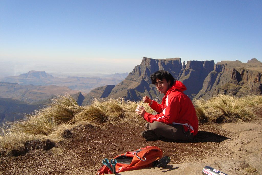 A man in a red shirt eats out of a tin cup on top of the Ampitheatre plateau in the Drakensberg mountains.